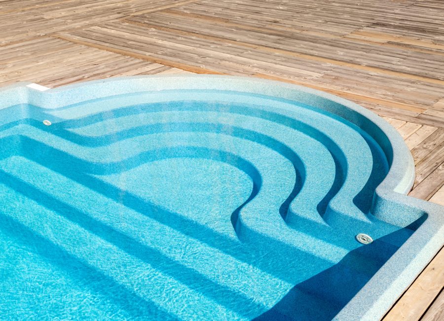 New modern fiberglass plastic swimming pool entrance step with clean fresh refreshing blue water on bright hot summer day at yard or resort hotel spa area. Wooden flooring deck of teak or larch board.